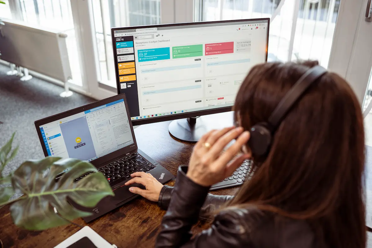 Mitarbeiterin von globboard arbeitet am Computer mit zwei Bildschirmen und nutzt ein Rezeptionscockpit-Dashboard. Auf dem Bildschirm sind Buchungs- und Anfragen-Statistiken sichtbar, während die Person mit Headset im Büro arbeitet.