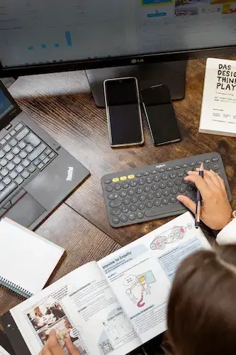 Eine Person arbeitet an einem Schreibtisch mit zwei Smartphones, einem Laptop und einer Tastatur. Auf dem Tisch liegt ein Buch mit technischen Diagrammen und ein Notizbuch.